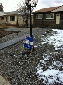 A Rahim Jaffer sign leaning gently against a lamp-post in a stone-filled yard