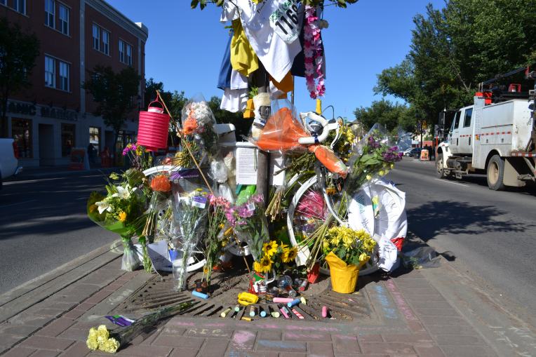 A white painted bicycle has been placed in a median on Whyte Ave. It is covered in flowers and cards for Isaak. A truck passes to the right.
