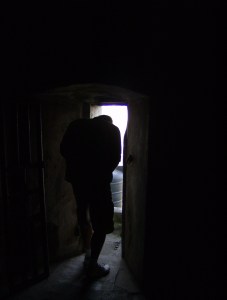 A man crouches to squeeze through a doorway out of a dark room and into the light.