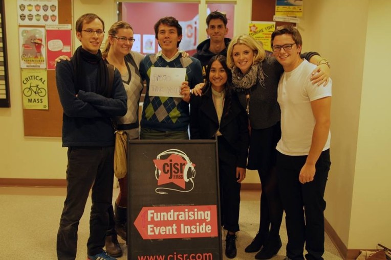 A group of radio hoodlums hug around a sign with the fundraising total from their FunDrive show. No windows are present, because CJSR is forever cursed to live in the basement.